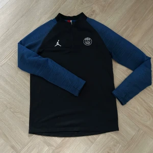 PSG x Jordan långärmad träningströja - Snygg svart träningströja från PSG och Jordan med marinblå ärmar och halv dragkedja. Tröjan har klubbmärke och Jumpman-logga på bröstet. Perfekt för fotbollsträning eller sportiga dagar.