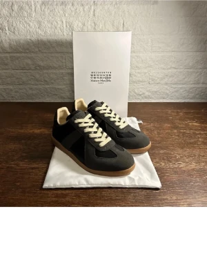 Maison Margiela svarta sneakers - Maison Margiela sneakers i svart med beige snörning och gummisula. Klassisk låg modell med rund tå och platt sula. Materialmix av mocka och skinn ger en lyxig känsla. Perfekt för dig som gillar stilrena och tidlösa sneakers. Priset kan diskuteras!!!
