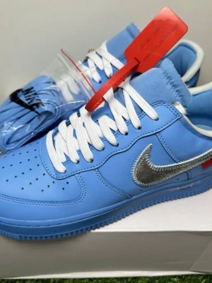 Nike Air Force 1 Off-White blå sneakers - Säljer ett par Nike Air Force 1 Off-White sneakers i ljusblått med vit snörning och en metallic silver swoosh. Skorna har en klassisk rund tå och platt sula, samt en röd tag-detalj. Materialet är skinn och syntet. Perfekt för dig som vill sticka ut med en unik färg. Finns i alla storlekar 