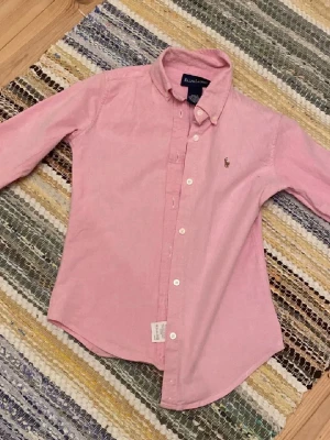 Rosa skjorta från Ralph Lauren - Säljer en klassisk rosa skjorta från Ralph Lauren med lång ärm och knappar framtill. Skjortan har en snygg krage och den ikoniska broderade loggan på bröstet. Materialet är mjuk bomull med lite stretch för extra komfort. Perfekt till jeans eller chinos.