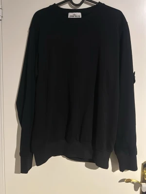 Svart långärmad tröja Stone Island - Svart långärmad tröja från Stone Island med klassisk rund hals och ribbade muddar. Tröjan har den ikoniska Stone Island-loggan på vänster ärm, fäst med två knappar. Materialet känns mjukt och är troligtvis bomull. Perfekt för dig som gillar streetwear och stilrena plagg.