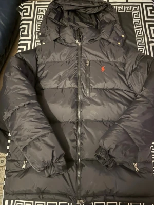 Svart pufferjacka från Polo Ralph Lauren - Svart pufferjacka från Polo Ralph Lauren med röd logga på bröstet. Jackan har huva, flera dragkedjor och en quiltad design som ger en schysst volym. Materialet är syntet och polyester, perfekt för kalla dagar. Snygg och stilren look med praktiska fickor.    Pris kan diskuteras? Notera att storleken känns som S !!    