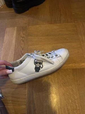 Karl Lagerfeld vita sneakers med tryck - Säljer ett par vita sneakers från Karl Lagerfeld med coolt tryck av Karl på sidan och svart detalj på hälen. Skorna har snörning, rund tå och är gjorda i skinn. Snygga och stilrena med logga på både sula och bakkappa.