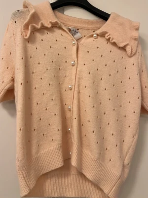 Beige kofta med pärlknappar och volangkrage - Supersöt beigerosa kofta från JS Millenium med korta ärmar, volangkrage och pärlknappar framtill. Stickad med små prickar i mönstret och ribbade muddar vid ärmslut och nederkant. Perfekt för dig som gillar gulliga detaljer och mjuka material.