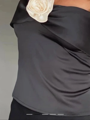 Svart bandeau tubtopp med rosdetalj (viral) - Säljer en svart bandeau tubtopp i glansigt satinmaterial. Toppen är axelbandslös och har en dekorativ ljusbeige ros framtill som ger en unik och trendig look. Perfekt för dig som vill sticka ut med en cool detalj. Passar dig som gillar stilrena plagg med twist. Blomman är lite mer grå än i bilderna. 
