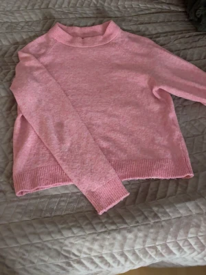 Rosa stickad tröja från Vero Moda - Mysig ljusrosa stickad tröja från Vero Moda med ribbade muddar vid hals, ärmslut och nederkant. Tröjan har en enkel, klassisk design med lång ärm och rundad hals. Perfekt för att addera lite färg till din garderob.