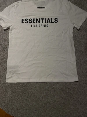 Vit Essentials Fear of God t-shirt - Vit t-shirt från Essentials Fear of God med svart tryck på ryggen. Klassisk rund hals och korta ärmar. Materialet är mjuk bomull och passformen är normal. Perfekt för dig som gillar cleana och stilrena streetwear-plagg.