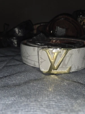 Vit Louis Vuitton bälte med guldspänne - Säljer ett stilrent vitt bälte från Louis Vuitton med klassiskt LV-spänne i guld. Bältet har ett diskret mönster med LV-loggor och är tillverkat i skinn. Perfekt för att lyfta din outfit med en lyxig touch.