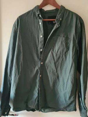 Grön skjorta med småmönster från Lager 157 - Säljer en grön långärmad skjorta från Lager 157 i storlek L. Skjortan har ett diskret småmönster i blått, klassisk krage, bröstficka och vita knappar framtill. 