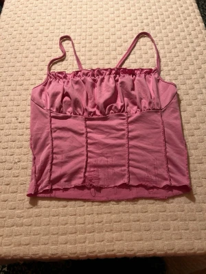 Rosa croppad linnetopp med smala axelband - Säljer en rosa croppad linnetopp i storlek S med smala axelband och rynkade detaljer upptill. Toppen har synliga sömmar och en mjuk, stretchig känsla. Perfekt för varma dagar och enkel att matcha med jeans eller kjol.