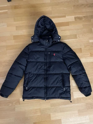 Svart pufferjacka från Polo Ralph Lauren - Svart pufferjacka från Polo Ralph Lauren med huva och dragkedjor på fickorna. Jackan har ett rött Polo-logo broderat på bröstet och är tillverkad i polyester. Perfekt för kalla dagar med sin tjocka och quiltade design. Fråga om du undrar nått