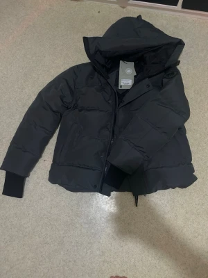 Svart pufferjacka från Canada Goose - Säljer en svart pufferjacka från Canada Goose, modell Fusion. Jackan har huva, ribbade muddar och dragkedja framtill. Perfekt för kalla dagar och riktigt snygg till streetwear-looken.