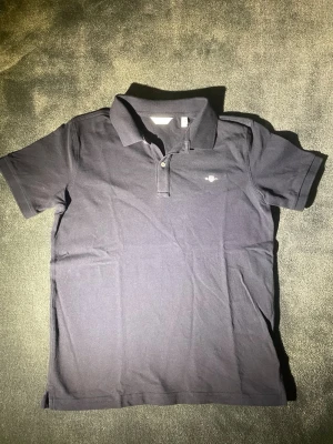 Mörkblå pikétröja från GANT - Säljer en svart pikétröja från GANT med klassisk krage och två knappar framtill. Tröjan har korta ärmar och en liten broderad logga på bröstet. Materialet är mjuk bomull som känns skönt mot huden. Perfekt för en chill och stilren look.