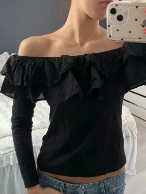 Svart offshoulder topp med volang H&M - Svart offshoulder topp från H&M i storlek S. Toppen har långa ärmar och bred volang runt hela axelpartiet som ger en snygg och trendig look. Perfekt att styla med jeans eller kjol för en cool vibe.