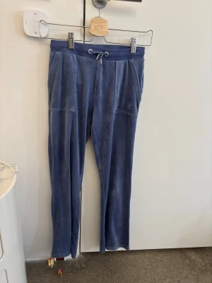 Blå mjukisbyxor från juicy couture med snörning - Säljer ett par blå mjukisbyxor i mjukt material med snörning i midjan och stora fickor fram. Baktill finns två fickor med knappar. Perfekta för chill dagar och har en relaxed passform. Köptes för 800kr 