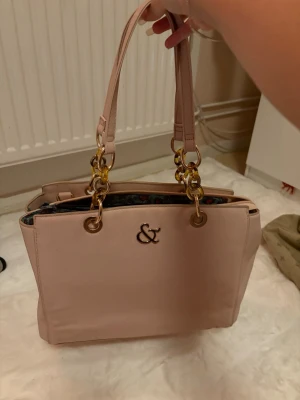 Ljusrosa handväska  - Säljer en ljusrosa handväska från & Other Stories med guldfärgade detaljer och dubbla handtag. Insidan har ett coolt leopardmönstrat foder i grönt och rött samt flera fack med dragkedja. Väskan är rymlig och perfekt för dig som vill ha både stil och funktion.