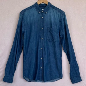 Blå jeansskjorta i S slim fit. Cubus - •Säljer en klassisk blå jeansskjorta i slim fit. •Skjortan har lång ärm, bröstficka och knappar hela vägen fram. •Har en viss (ombre-effekt), dvs. överdelen av skjortan är lite ljusare än resten. •Tillverkad i 100% bomull för en skön känsla och snygg tvättad look.                                         •Perfekt till jeans eller chinos.                        •Tvättad men har aldrig blivit använd.                 •ca-mått i cm: Axel: 42, Bröst: 46, Längd: 75