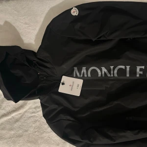 Svart vindjacka från Moncler - Säljer en svart vindjacka från Moncler med stor logga framtill och huva. Jackan har dragkedja, diskret Moncler-märke på ärmen och snygga detaljer som gör den riktigt clean. Perfekt för dig som vill ha en stilren och funktionell jacka.