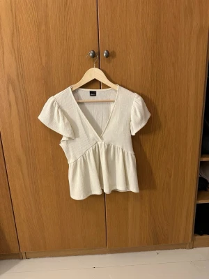 Vit ribbad omlottopp. - Säljer en söt vit ribbad topp från Gina Tricot i storlek S. Toppen har omlottdesign med v-ringning, korta ärmar och volang nedtill. 