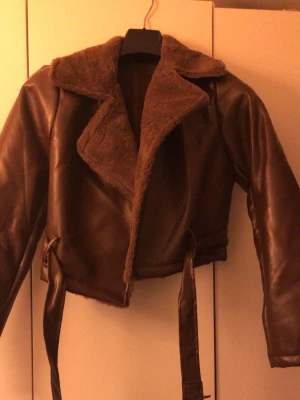 Brun teddyjacka med bälte - Säljer en brun teddyjacka med croppad passform och bred krage i mjuk pälsimitation. Jackan har ett snyggt bälte i midjan och är tillverkad i syntetmaterial med pälsdetaljer. Perfekt för dig som vill ha en cool och varm look.