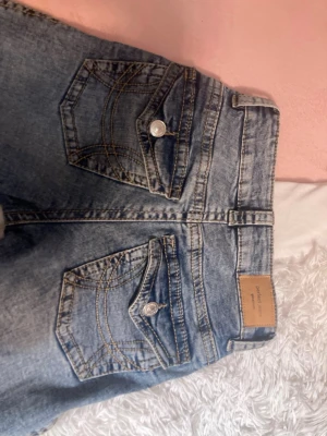 Perfekt Jeans ljusblå bootcut 34 - Säljer ett par ljusblå jeans från Perfect Jeans i storlek 34. Modellen har bootcut-ben med rå kant nedtill och klassiska fem fickor, varav bakfickorna har lock och knapp. Jeansen har normal passform och är tillverkade i jeansmaterial med snygga kontrastsömmar.