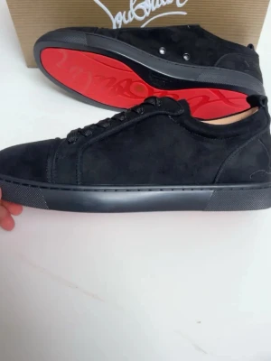 Svarta sneakers från Christian Louboutin - Säljer ett par svarta sneakers från Christian Louboutin med ikonisk röd sula och broderad logga på hälen. Skorna är tillverkade i mocka och har snörning framtill. Snygg och stilren design som sticker ut med den röda undersidan.