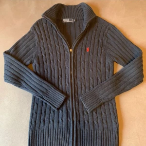 Mörkblå stickad kofta från Polo Ralph Lauren - Säljer en mörkblå stickad kofta från Polo Ralph Lauren med dragkedja och hög krage. Klassisk kabelstickad design och röd broderad logga på bröstet. Perfekt för lager-på-lager och snygg till vad som.