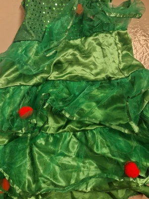 Grön julklänning med volanger - Unik grön klänning med flera lager glansigt tyg och volanger, dekorerad med röda bollar och paljetter upptill. Klänningen har en festlig look som påminner om en julgran och är tillverkad i syntetmaterial med transparenta detaljer. Ingen hatt ingår. För små barn one size