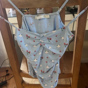 Blommig ljusblå linnetopp med rynk - Supersöt ljusblå linnetopp från 157 med smala axelband och rynkdetalj framtill. Toppen har ett blommigt mönster med små vita och röda blommor över hela tyget. Perfekt för varma dagar och enkel att matcha med jeans eller kjol.