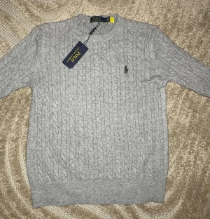 Grå kabelstickad tröja Polo Ralph Lauren - Säljer en grå kabelstickad tröja från Polo Ralph Lauren. Tröjan har rund hals, lång ärm och den klassiska broderade loggan på bröstet. Tillverkad i mjuk bomull och har en clean, tidlös look som funkar till allt.