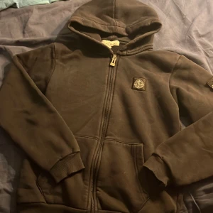 Svart hoodie från Stone Island - Svart hoodie från Stone Island med ikoniska kompasspatchen på ärmen och bröstet. Tröjan har huva, dragkedja framtill och ribbade muddar. Tillverkad i mjuk bomull, perfekt för chill dagar. 