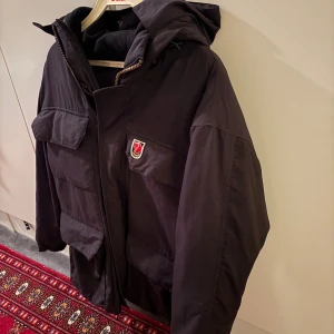 Svart parkajacka från Fjällräven XS - Svart parkajacka från Fjällräven med huva och stora fickor framtill. Jackan har dragkedja och knappar, samt ett klassiskt Fjällräven-märke på bröstet. Materialet är slitstark och jackan är fodrad med mjuk fleece på insidan för extra värme. Använts en del därav det låga priset. Men den har många år kvar att ge!