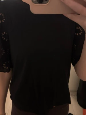 Tröja - Säljer en svart blus från dobbie med rund hals och korta ärmar. Ärmarna har broderade blommönster och håldetaljer som ger en cool och unik look. Blusen är gjord i mjuk bomull och har en normal passform. Perfekt att styla med jeans eller kjol.