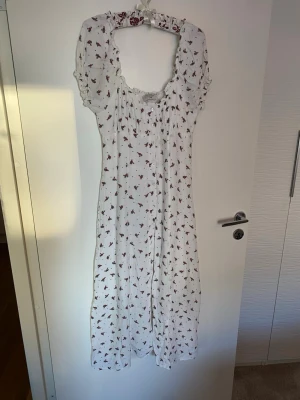 Superfin sommarklänning med fin urringning - Superfin vit långklänning med små röda blommor och gröna blad. Klänningen har puffiga korta ärmar, knappar framtill och en söt rynkad kant vid halsen. Klänningen har knappar hela vägen och lent innerfoder. Materialet är lätt och luftigt, perfwkt till sommaren eller till fest. Passformen är normal och klänningen har en romantisk vibe. En absolut favorit, men tyvärr för liten för mig. Storleken överensstämmer inte med storleken på bilden XL, det är en Medium. 