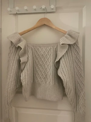 Beige stickad tröja med volang H&M - Supersnygg beige stickad tröja från H&M i storlek XS. Tröjan har shoulder-design med breda volanger längs axlarna och ett flätstickat mönster både fram och bak köpt på hm för flera år sedan för ca 450kr knappt använd 
