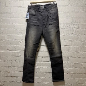 Versuchkind Berlin drop crotch slim fit - Säljer ett par grå jeans från VERSUCHSKIND Berlin med slitna detaljer och tvättad look. Byxorna har raka ben, fem fickor och coola trasiga partier framtill. Materialet är robust denim och färgen är mörkgrå med ljusare partier. Perfekt för dig som gillar streetstyle.