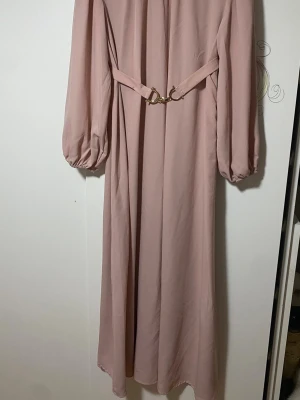 Ljusrosa långklänning med bälte - Säljer en elegant ljusrosa långklänning med puffiga långärmar och ett snyggt midjebälte med guldfärgat spänne. Klänningen har en mjuk, flowy passform och är tillverkad i ett lätt syntetmaterial som faller fint. 