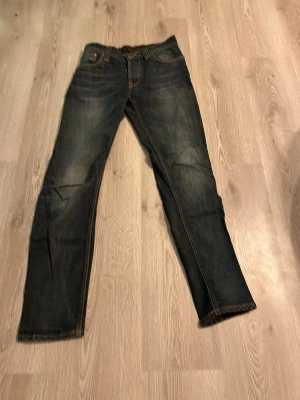 Mörkblå Nudie Jeans, W29 L32 - Säljer ett par mörkblå jeans från Nudie Jeans med klassisk femficksdesign och kontrastsömmar. Modellen har raka ben och normal passform, tillverkade i slitstarkt bomullsmaterial. Snygga detaljer på bakfickorna och läderpatch med logga bak i midjan.