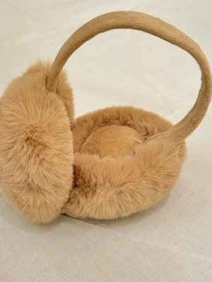Beige fluffiga öronmuffar i fuskpäls - Supersöta och mjuka öronmuffar i beige fuskpäls. De har en rund form och bred båge som sitter bekvämt över huvudet. Perfekta för att hålla öronen varma under kalla dagar och ger en mysig vinterlook.