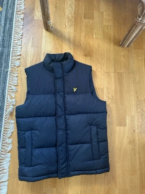 Mörkblå dunväst från Lyle & Scott - Snygg mörkblå dunväst från Lyle & Scott med gul logga på bröstet. Västen har hög krage, två fickor framtill och quiltad design. Perfekt att slänga över en hoodie eller tröja för extra värme.