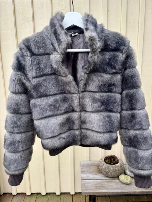 Grå pälsjacka i storlek 34 - Säljer en fluffig och riktigt snygg grå pälsjacka med horisontella paneler. Jackan har hög krage, långa ärmar och ribbade muddar vid ärmslut och nederkant. Perfekt för dig som vill ha en statementjacka i vinter. 