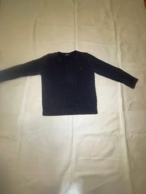 Mörkblå stickad tröja från Polo Ralph Lauren - Säljer en mörkblå stickad tröja från Polo Ralph Lauren med klassiskt kabelstickat mönster och röd broderad logga på bröstet. Tröjan har rund hals, ribbade muddar och långa ärmar. Perfekt för dig som gillar stilrena och tidlösa plagg.