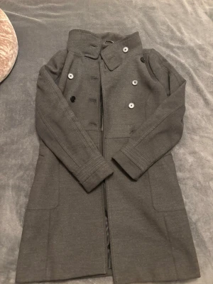 Grå kappa från Zara Trafaluc🫶🏻 - Säljer en stilren grå kappa från Zara Trafaluc i storlek L. Kappan har dubbelknäppning med svarta och grå knappar, klassisk krage och två stora fickor framtill. Materialet är mjukt och känns som ull, perfekt för kyliga dagar. Ens knappen har trillat av, den finns kvar men har bara inte haft orken att sy på den. Jag kommer skicka med den men kan tänka mig att sy på den om den säljs🤍