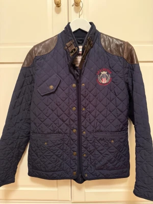 Mörkblå quiltad jacka Lexington - Quiltad mörkblå jacka från Lexington med bruna skinnpartier på axlarna och broderat emblem på bröstet. Jackan har snygga detaljer som knappar, fickor och spänne vid halsen. 