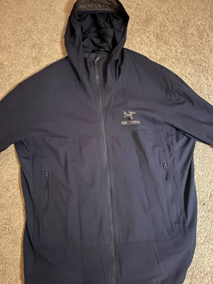 Mörkblå vindjacka från Arc'teryx - Säljer en mörkblå vindjacka från Arc'teryx med huva och hel dragkedja framtill. Jackan har två sidofickor med dragkedja och en stilren logga på bröstet. Perfekt för dig som vill ha en lätt och funktionell jacka till vardags.