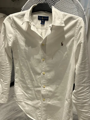 Vit skjorta från Ralph Lauren - Klassisk vit skjorta från Ralph Lauren med broderad logga på bröstet. Skjortan har lång ärm, knappar framtill och är gjord i mjuk bomull. Perfekt till jeans eller chinos för en clean och stilren look. Storlek L i barn storlek som motsvarar ungefär 164-170
