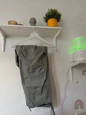 Grå täckbyxor med snörning- oanvänt  - Säljer ett par grå täckbyxor med elastisk midja och snörning vid bensluten. Byxorna har en loose passform och är gjorda i ett syntetmaterial som passar perfekt när du vill hålla dig varm. Snyggt och praktiskt val för kalla dagar.