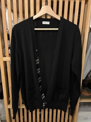 Svart kofta från Dries Van Noten - Svart kofta från Dries Van Noten i storlek medium. Koftan har lång ärm, två framfickor och knäppning med knappar. Insidan av knappslån har ett randigt tyg i svart, vitt och rött som ger en snygg detalj. Materialet känns mjukt och är stickat.