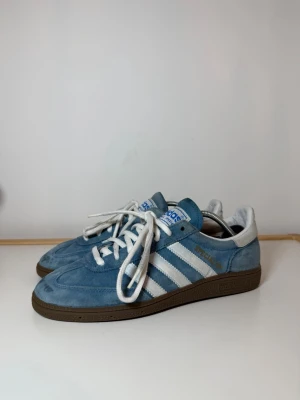 Adidas Spezial blå sneakers - Säljer ett par Adidas Spezial sneakers i blå mocka med vita detaljer och klassiska tre ränder på sidan. Skorna har vit snörning, brun platt sula och Adidas-logga i blått på tungan och hälen. Perfekt för dig som gillar retrostil och streetwear.