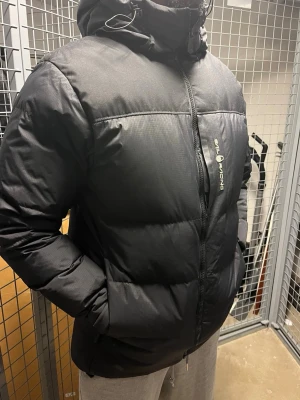 Svart Sail Racing dunjacka med huva - Säljer en svart dunjacka från Sail Racing med huva och dragkedja framtill. Jackan har en clean look med logga på bröstet och patch på ärmen. Flera fickor och justerbar huva med snörning. Perfekt för kalla dagar och riktigt snygg stil.